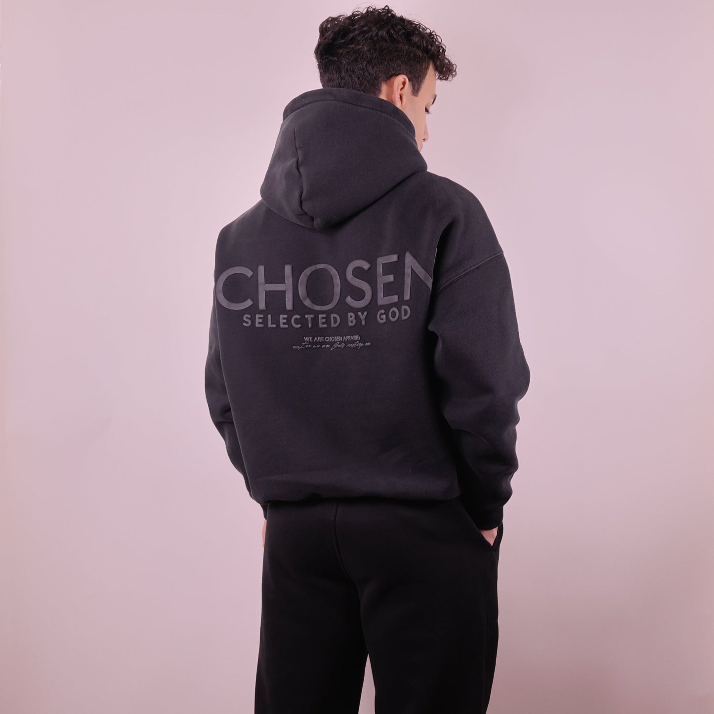 Person wearing a black hoodie with Chosen Selected by God text on the back against a light pink background