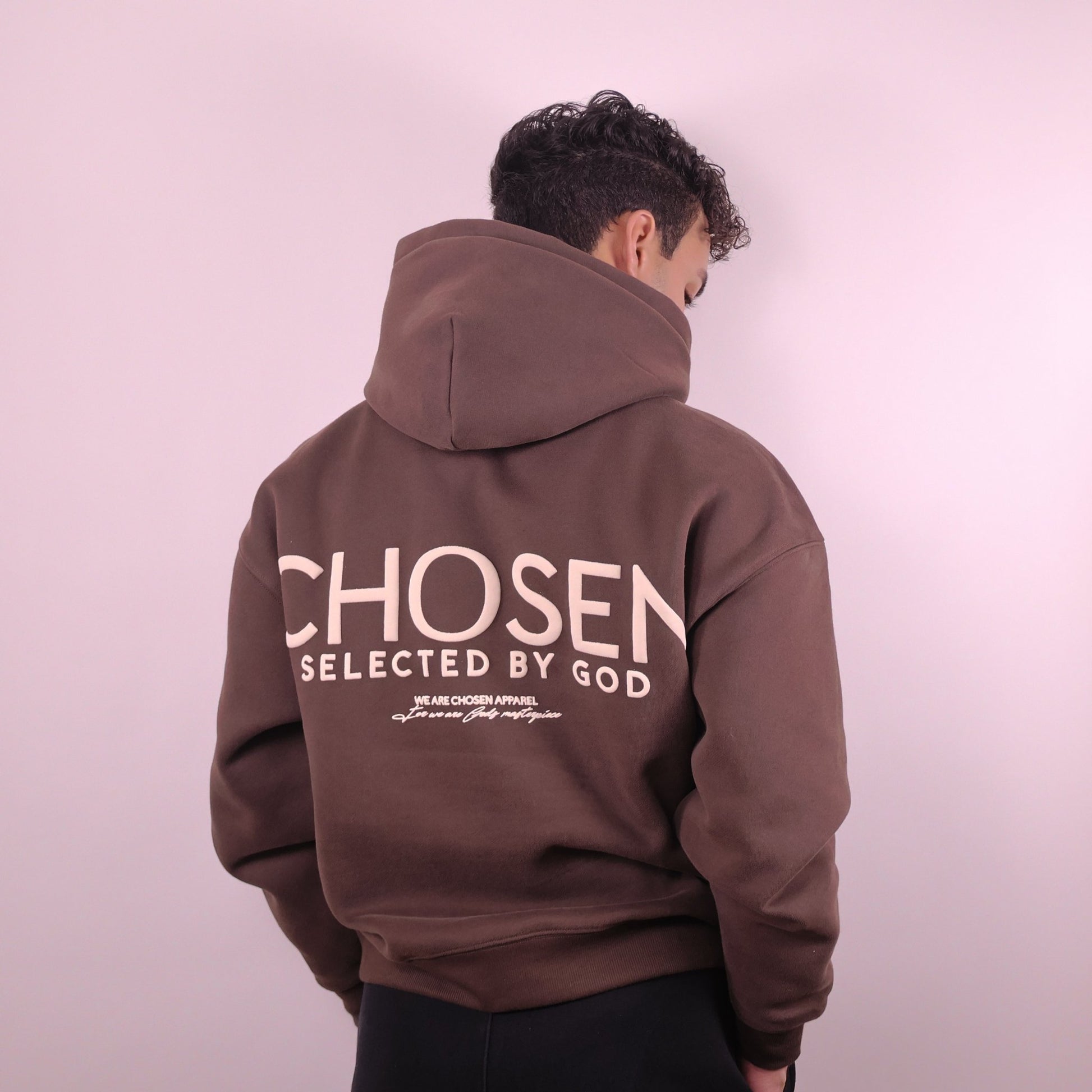 Person wearing a brown hoodie with Chosen Selected by God text on the back against a light pink background