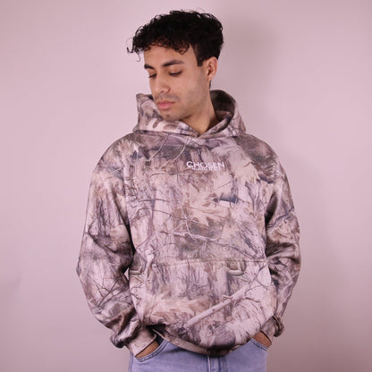 Person wearing a camouflage hoodie with a brand logo on a plain background