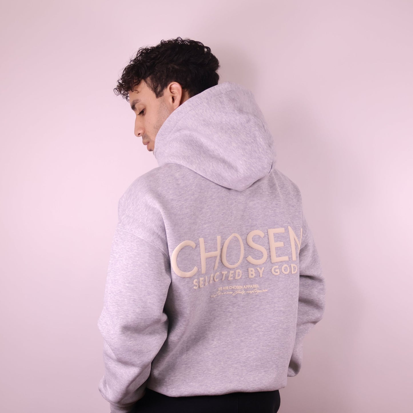 Person wearing a gray hoodie with Chosen Selected by God text on the back against a light pink background