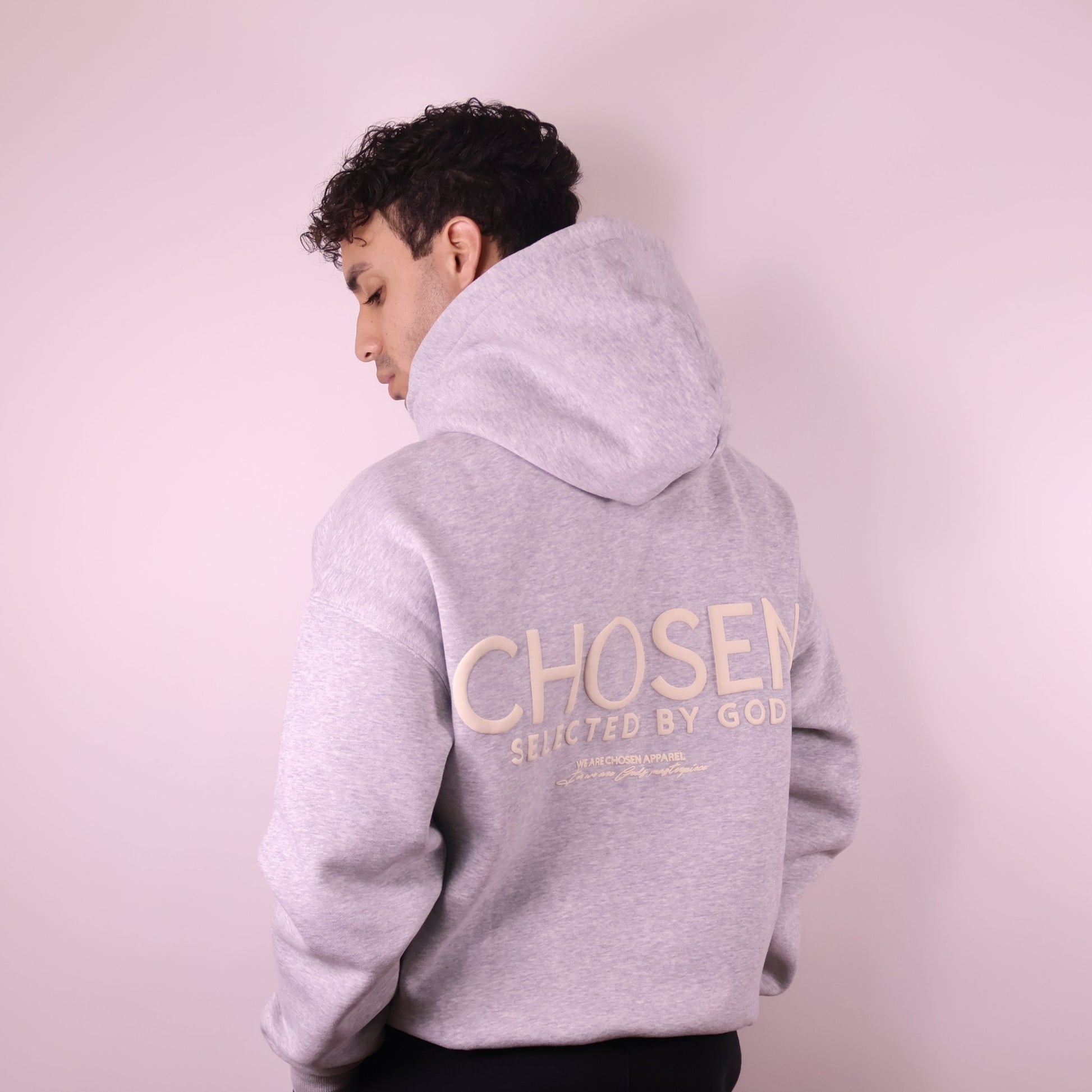 Person wearing a gray hoodie with Chosen Selected by God text on the back against a light pink background