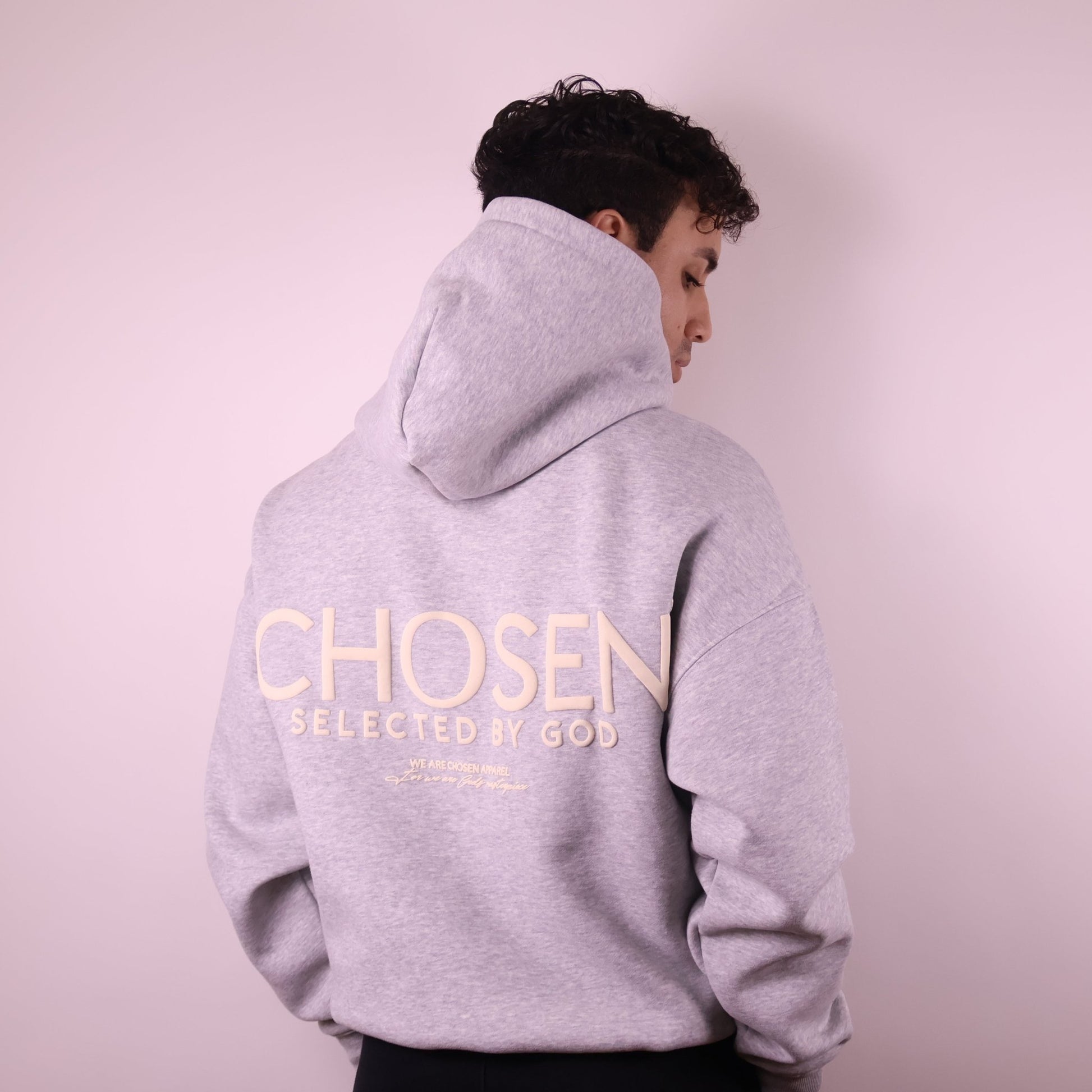 Person wearing a gray hoodie with Chosen Selected by God text on the back against a light pink background