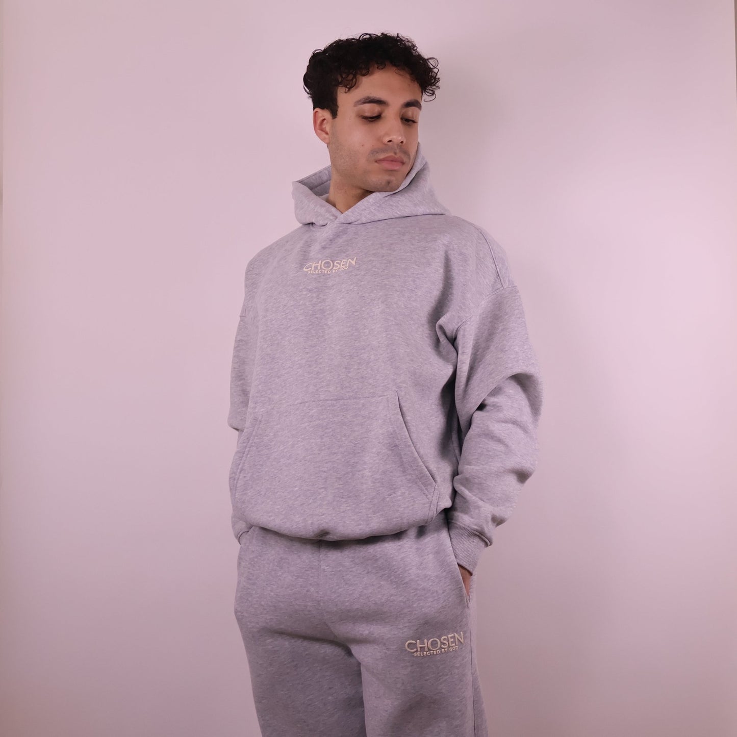 Person wearing a gray hoodie and pants with Chosen branding on a light pink background