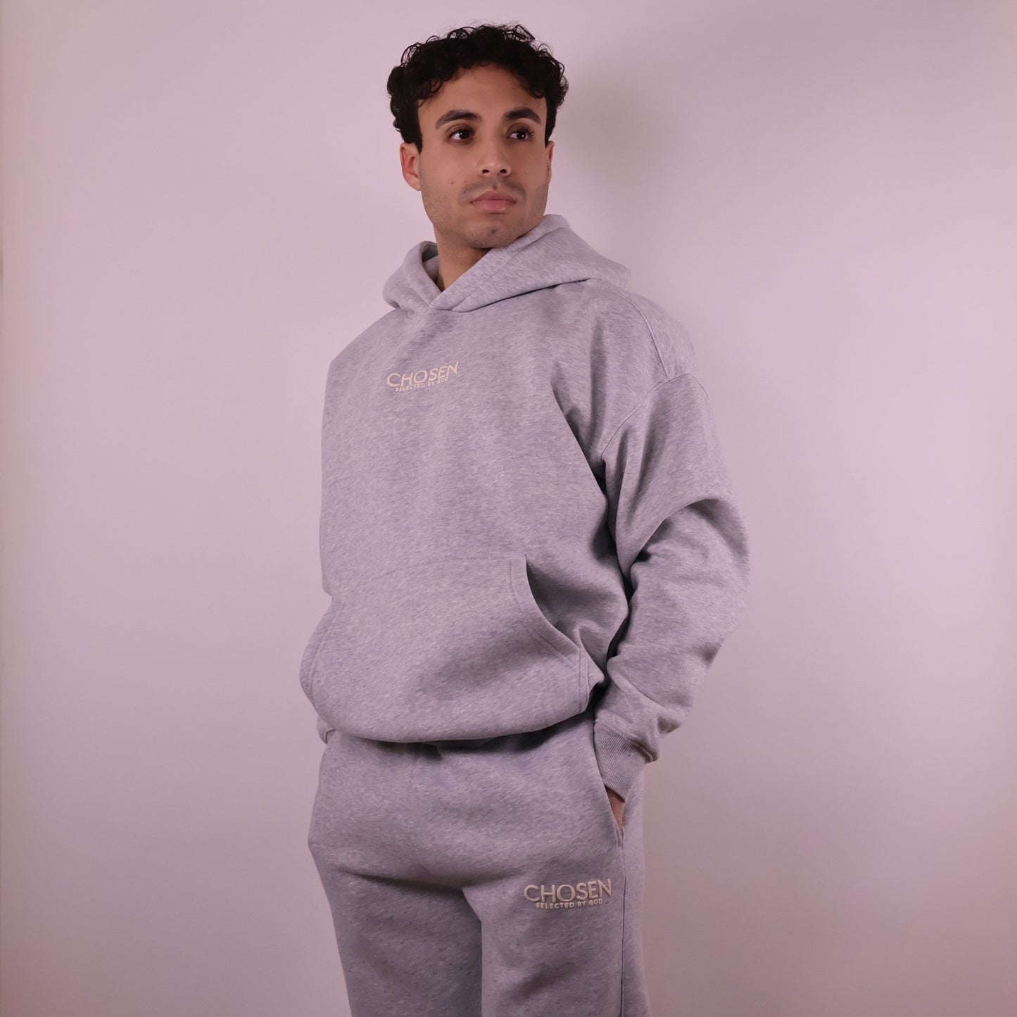 Person wearing a gray hoodie and pants with Chosen branding on a light pink background