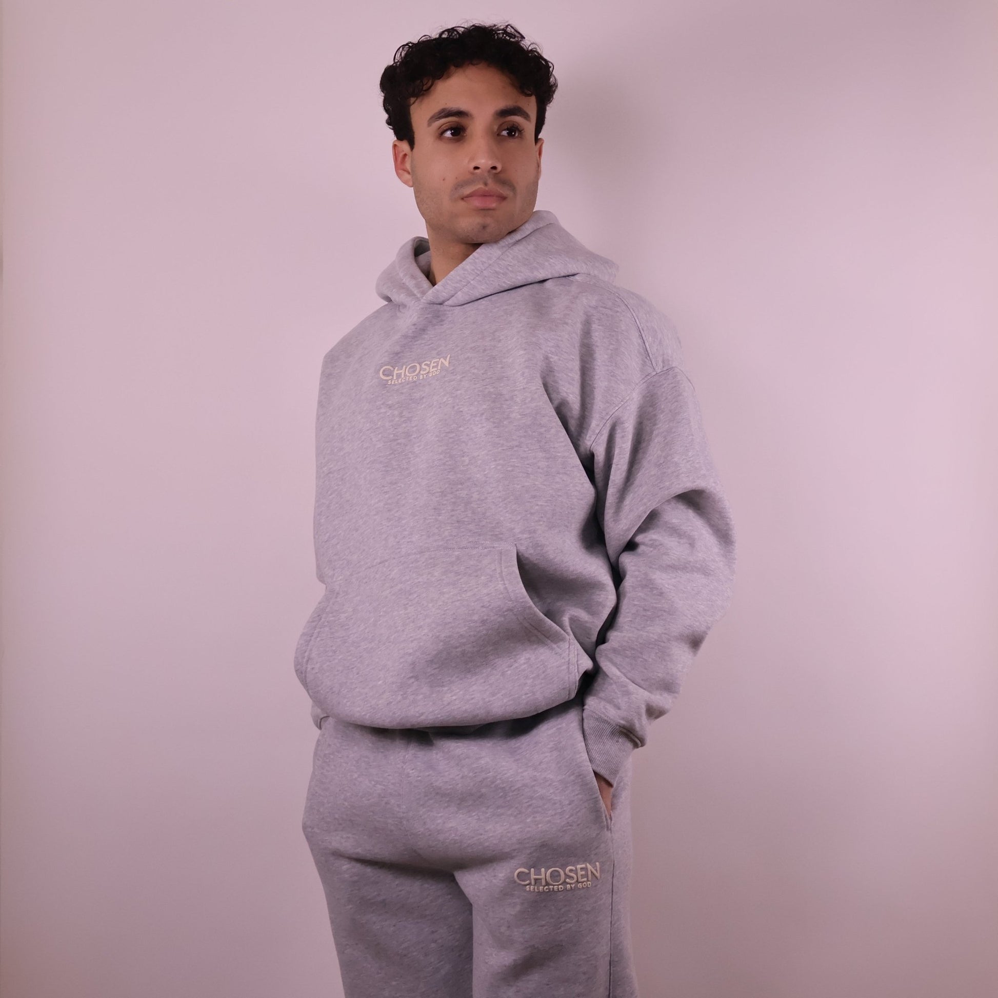 Person wearing a gray hoodie and pants with Chosen branding on a light pink background