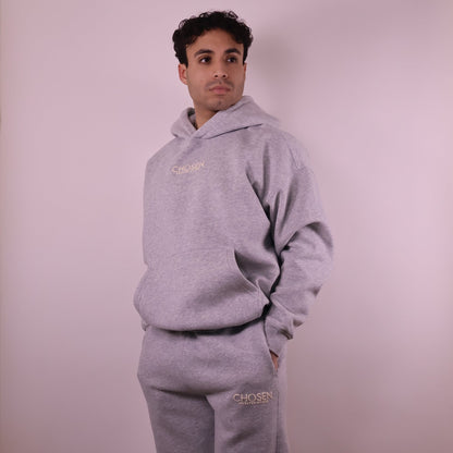 Person wearing a gray hoodie and pants with Chosen branding on a light pink background