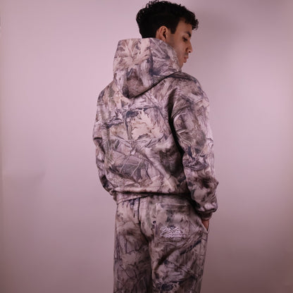 Person wearing a camouflage hoodie and pants against a plain background