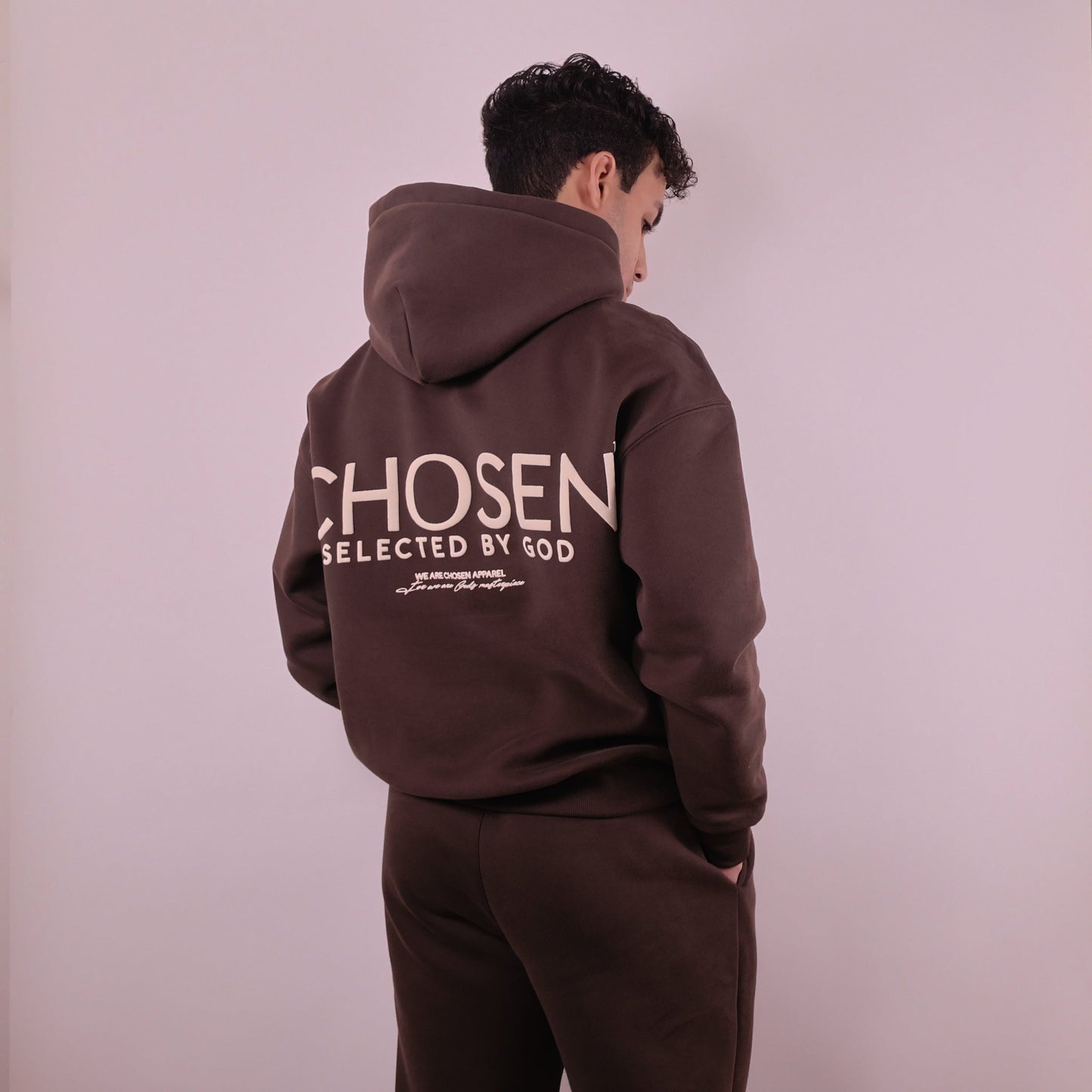 Person wearing a brown hoodie with Chosen Selected by God text on the back against a plain background